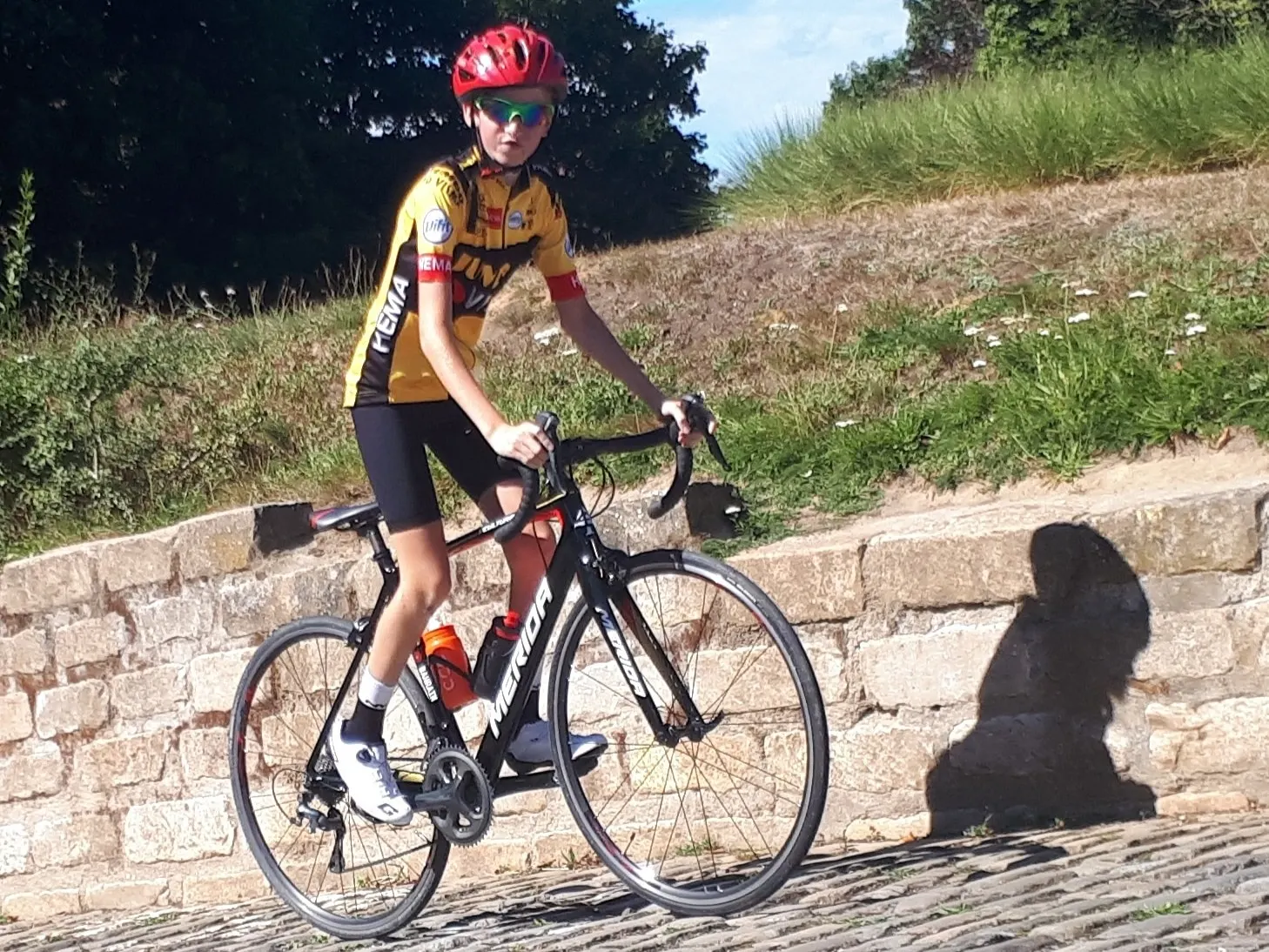 Young Xander in a Jumbo-Visma jersey on a black Merida road bike, beside a stone wall.
