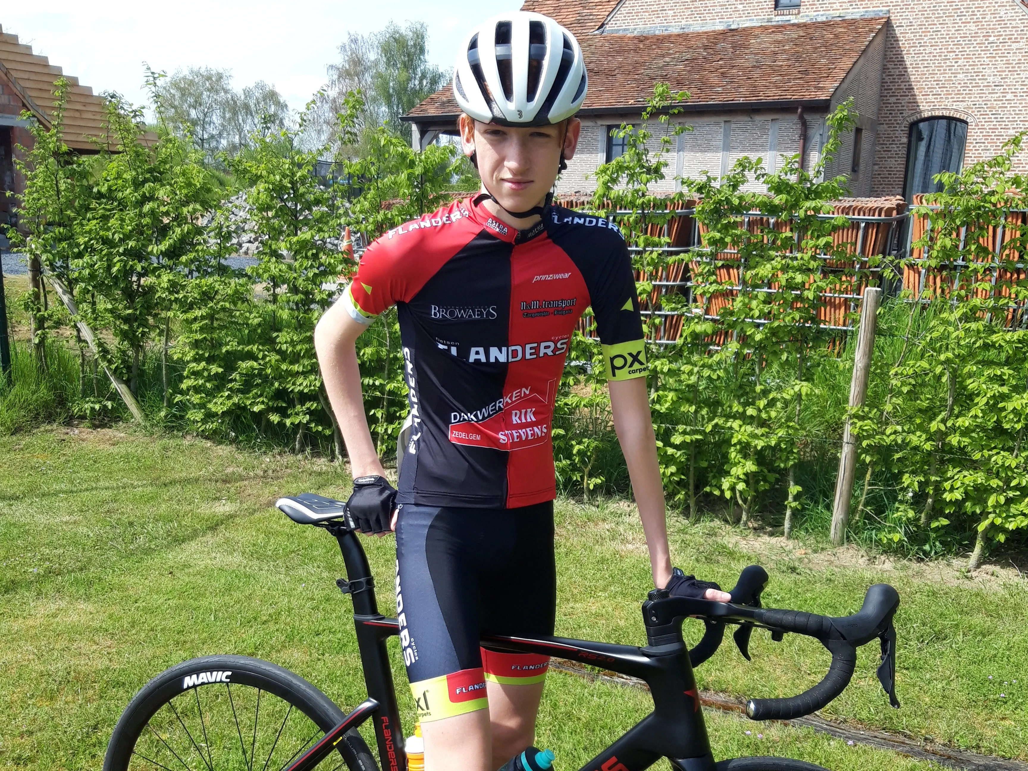 Xander in a red and black Flanders team kit, standing with his road bike in a garden.